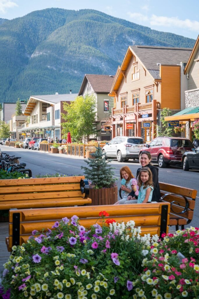 A colorful street scene in a mountain town. The image features a person on a bench with vibrant flowers in the foreground, with mountain views and charming local architecture in the background.