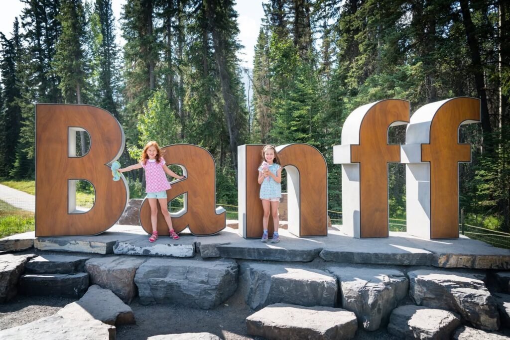 Visitors stand next to giant wooden letters spelling Banff on a stone base in a lush forest setting.