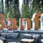 Visitors stand next to giant wooden letters spelling Banff on a stone base in a lush forest setting.
