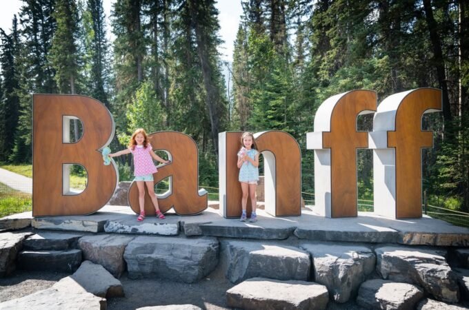 Visitors stand next to giant wooden letters spelling Banff on a stone base in a lush forest setting.