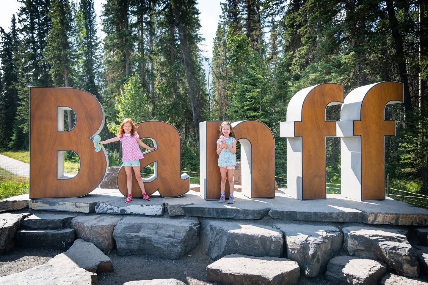 Bye Bye Banff Visitors stand next to giant wooden letters spelling Banff on a stone base in a lush forest setting.