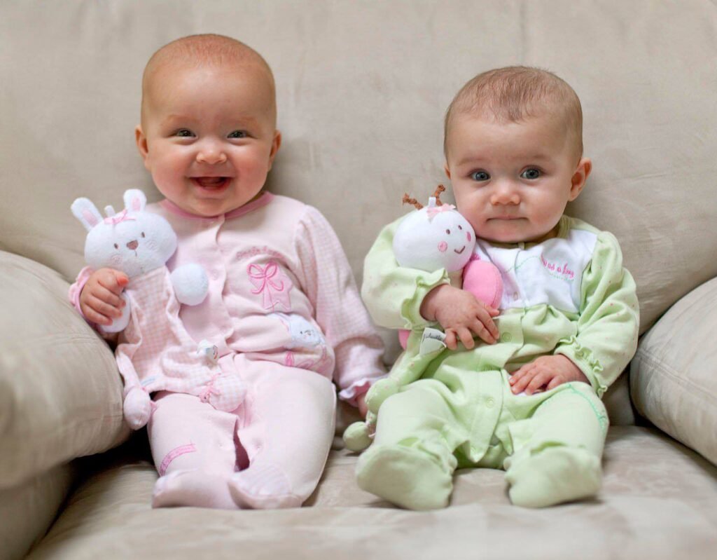 Two babies in matching pastel footed pajamas sit on a beige sofa holding plush stuffed animals.
