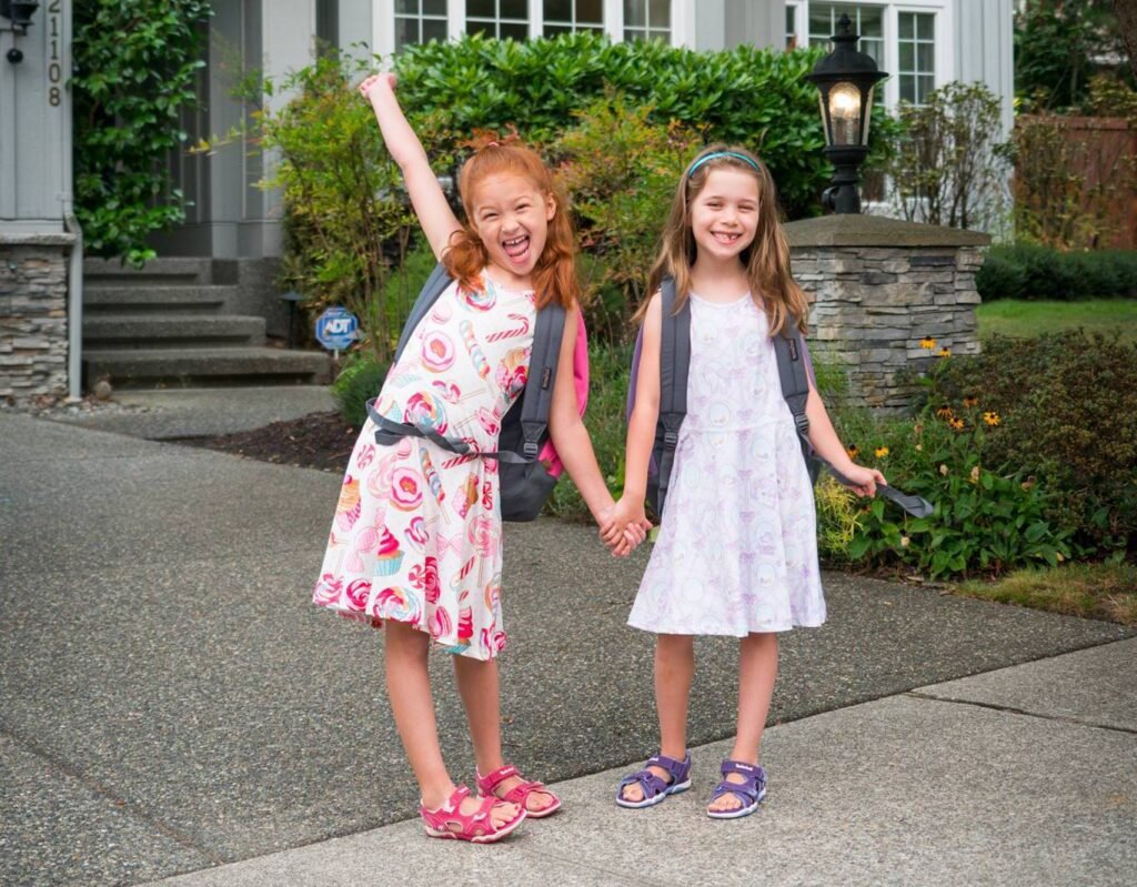 Two smiling children wearing backpacks, holding hands on a driveway in front of a house.