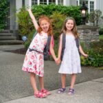 Two smiling children wearing backpacks, holding hands on a driveway in front of a house.