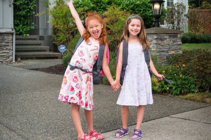 Two smiling children wearing backpacks, holding hands on a driveway in front of a house.