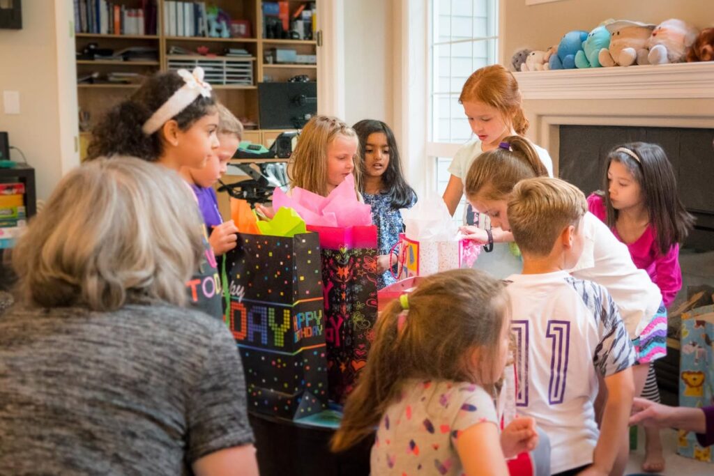 A group of children and adults enjoying a birthday party in a cozy living room. Gift bags and a fireplace provide a warm, festive setting.