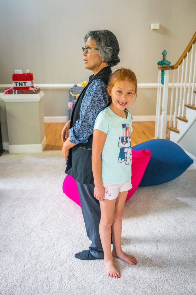 A grandparent and child smile while standing near playful bean bags in a bright home interior. Surrounding them are colorful cushions on a white carpet.