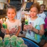 A group of children holding cupcakes with lit green candles at a party.