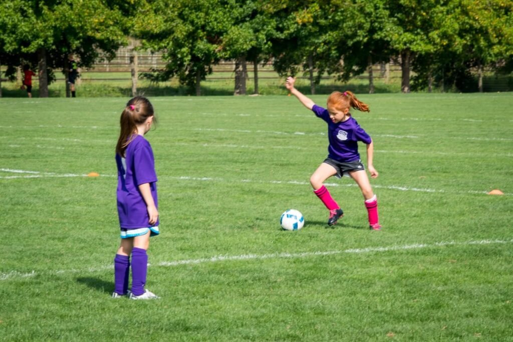 Two young players in purple jerseys on a grassy field, with one balancing a ball on a raised knee.