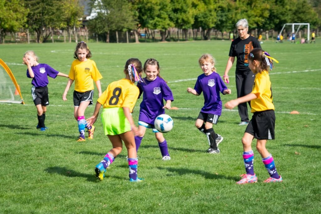 Young players in purple and yellow jerseys compete during a youth baseball game on a sunny field. A coach looks on as the young athletes move around the ball.