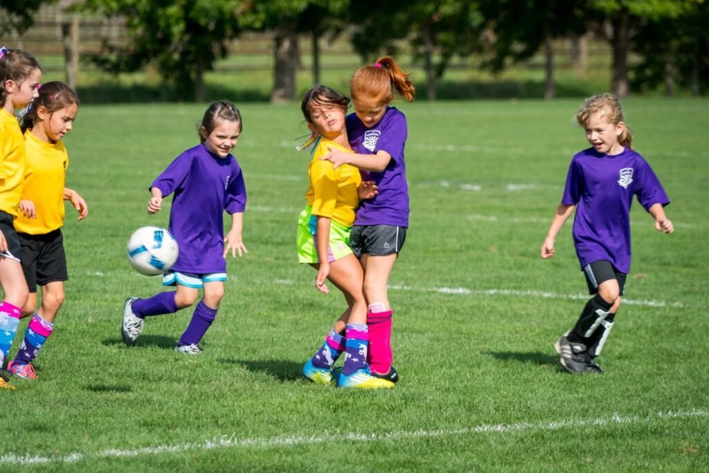 A group of young children in yellow and purple uniforms playing soccer on grass. Two players hug in the middle of the field during the game.