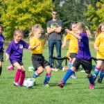 A group of children in purple and yellow uniforms play soccer on a field while an adult supervisor watches closely.