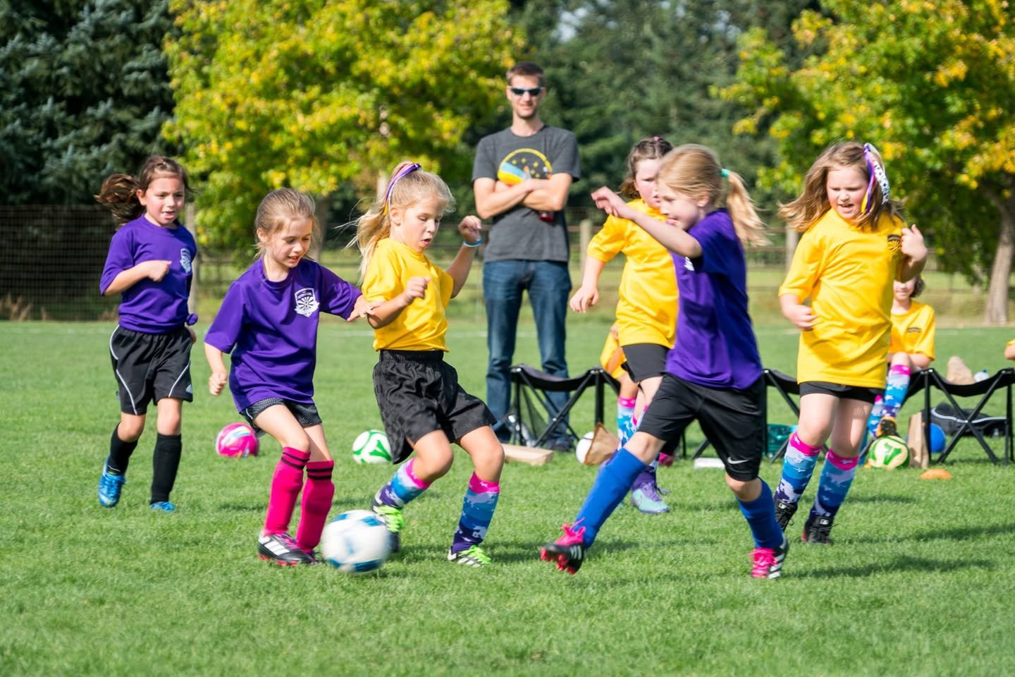 Cardinals vs. Unicorns A group of children in purple and yellow uniforms play soccer on a field while an adult supervisor watches closely.