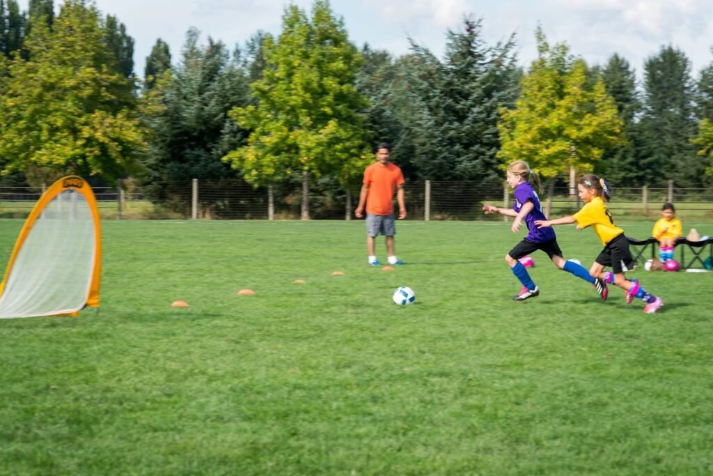 Children and an adult playing soccer on a grassy field in a park. The scene includes colorful uniform players, a portable goal, and trees in the background.