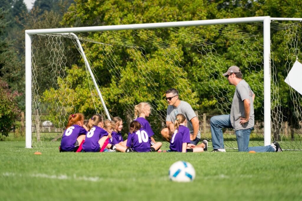 Young athletes in purple uniforms sit together on a soccer field while adults nearby observe, with a soccer goal and ball in the foreground.