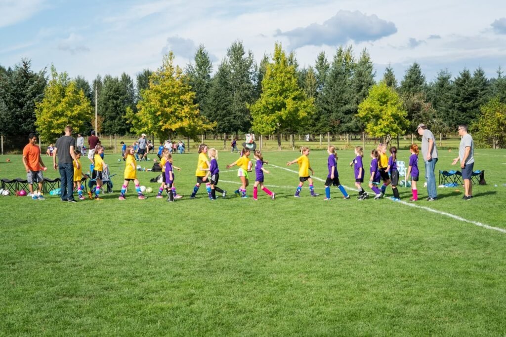 Young players in yellow and purple jerseys during a soccer practice on a grassy field with trees in the background.