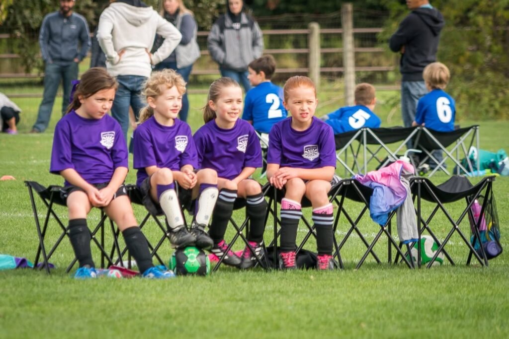 Young soccer players in purple uniforms sit on folding chairs at a local match. Teammates in blue jerseys stand nearby on the grass.
