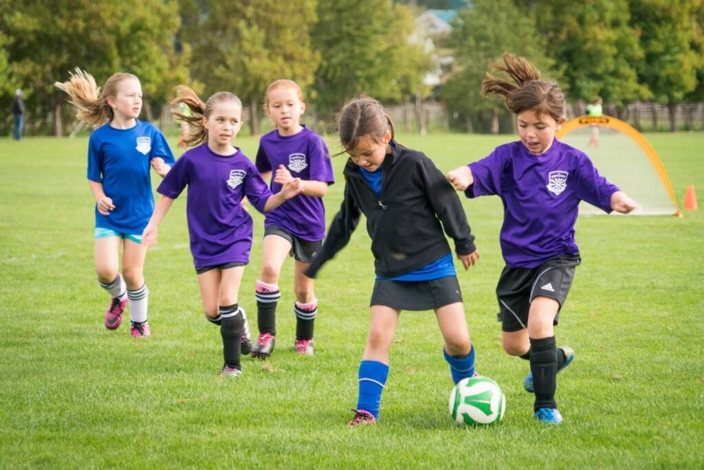 Children in purple, blue, and black uniforms play youth soccer on a bright green field.