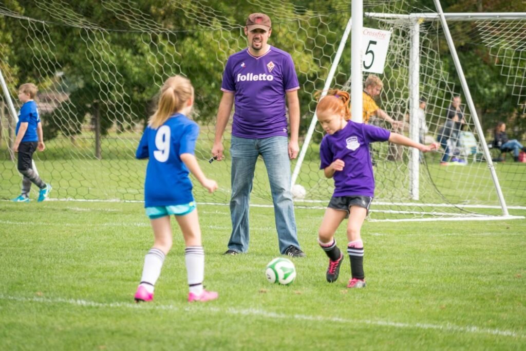 A group playing soccer on a sunny day, with a parent in a purple shirt watching the young players in blue and purple jerseys.