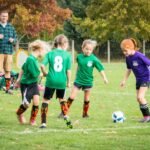 A group of children playing soccer in green and purple jerseys on a grass field in the Pacific Northwest.