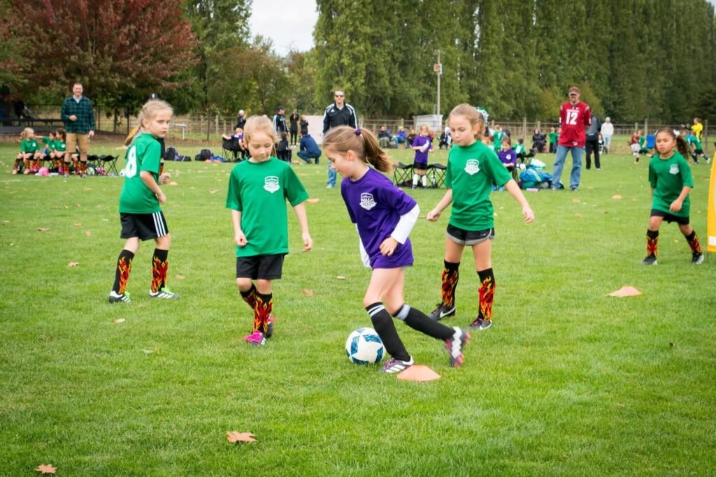 Children playing soccer on a grassy field wearing green and purple jerseys. One player in purple is kicking the ball.