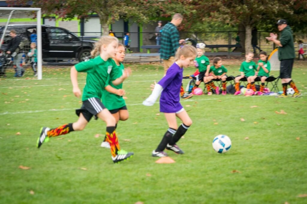 Young athletes competing in a lively soccer match, with a green and purple team. Teams of children cheer from the sidelines.