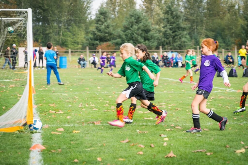 Young soccer players in green and purple jerseys compete on a grassy field. The action unfolds near the goal with the ball in play.