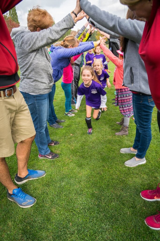 A group of children running through an outdoor archway formed by people holding bags tied on their heads, with one child in a purple shirt leading the line on a grassy field.