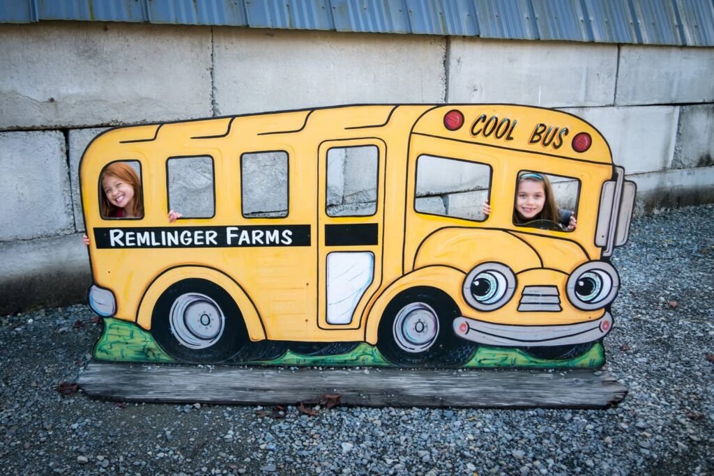 A large, colorful wooden cutout of a cartoon school bus at Remlinger Farms. Two visitors peek out of the windows, wearing smiles.