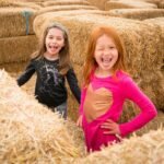 Two children smiling and playing in a large pile of hay bales at an autumn farm, enjoying the outdoors.