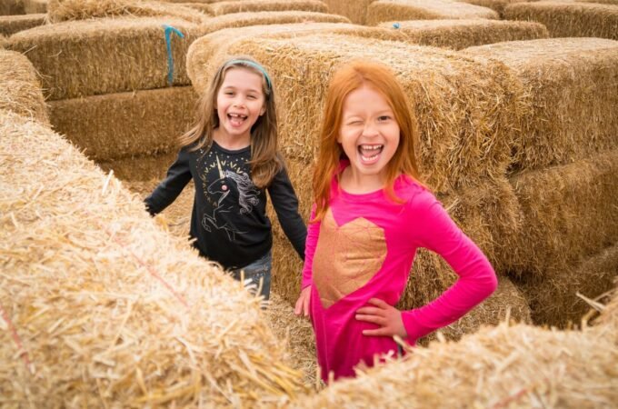 Two children smiling and playing in a large pile of hay bales at an autumn farm, enjoying the outdoors.