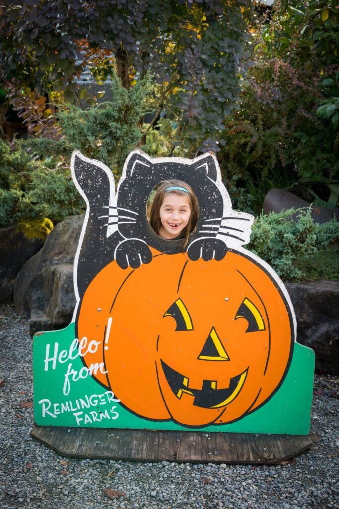 A cheerful child photographs through a cutout in a festive Halloween sign. The sign features a black cat climbing a large orange pumpkin with a sign reading 'Hello from Remlinger Farms.'