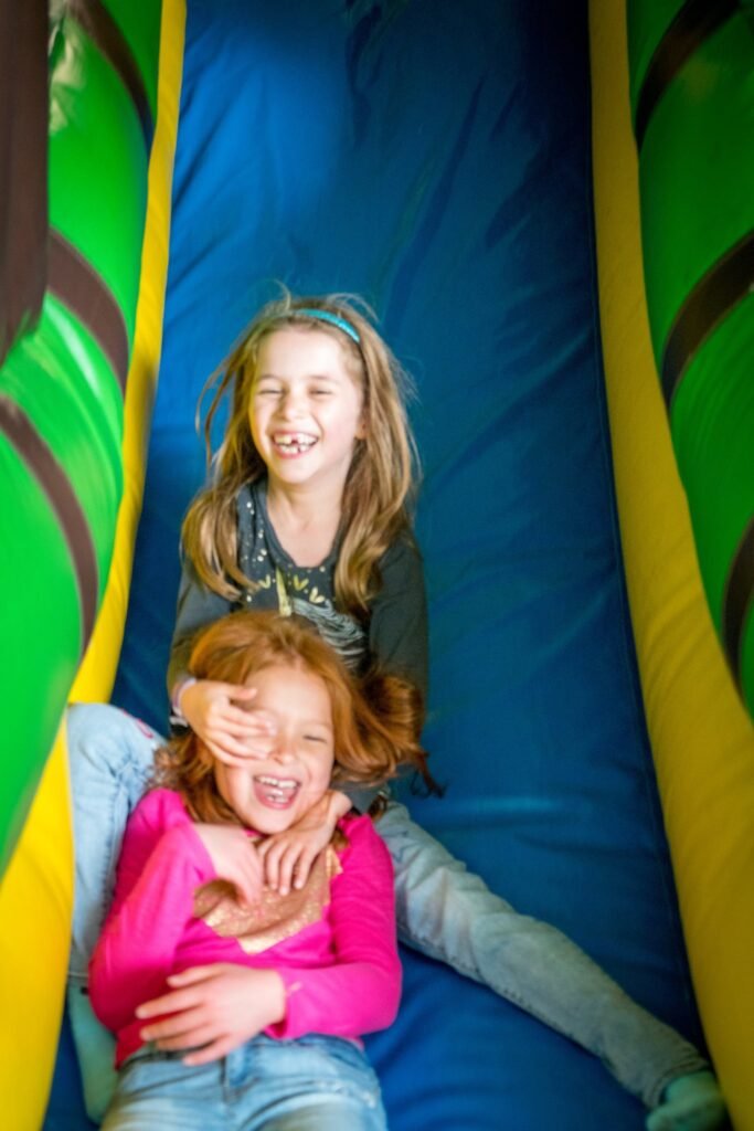 Multiple selfies of a girl in a pink shirt and another person sliding down a colorful inflatable slide at a family event.