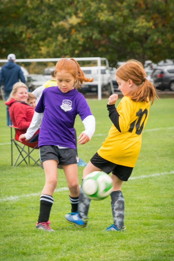 Two younger players with red hair running on a grass field to kick a soccer ball at a local fall match.