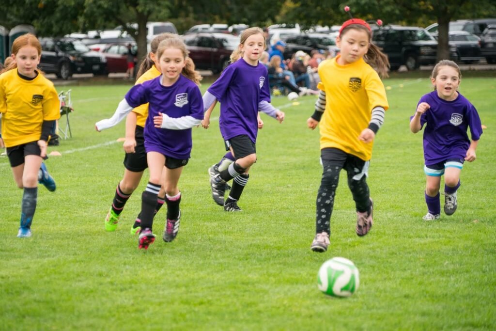 Children in yellow and purple jerseys chase a soccer ball on a grassy field.