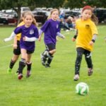 Children in yellow and purple jerseys chase a soccer ball on a grassy field.