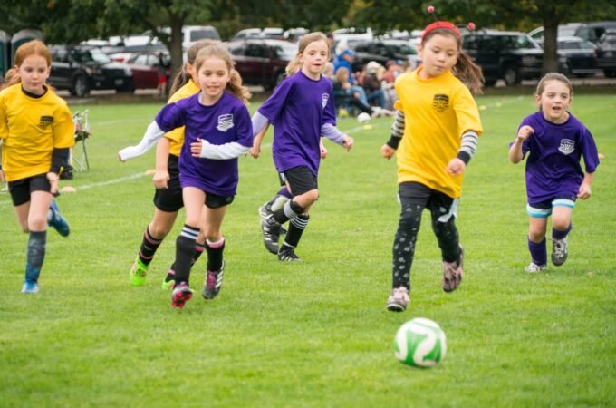 Children in yellow and purple jerseys chase a soccer ball on a grassy field.