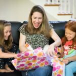 Three individuals enjoy wrapping colorful gifts together on a black leather couch, sharing smiles and laughter in a cozy home setting. The vibrant floral wrapping paper adds a festive touch to the heartwarming scene.