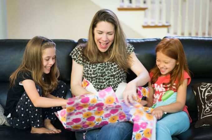 Three individuals enjoy wrapping colorful gifts together on a black leather couch, sharing smiles and laughter in a cozy home setting. The vibrant floral wrapping paper adds a festive touch to the heartwarming scene.