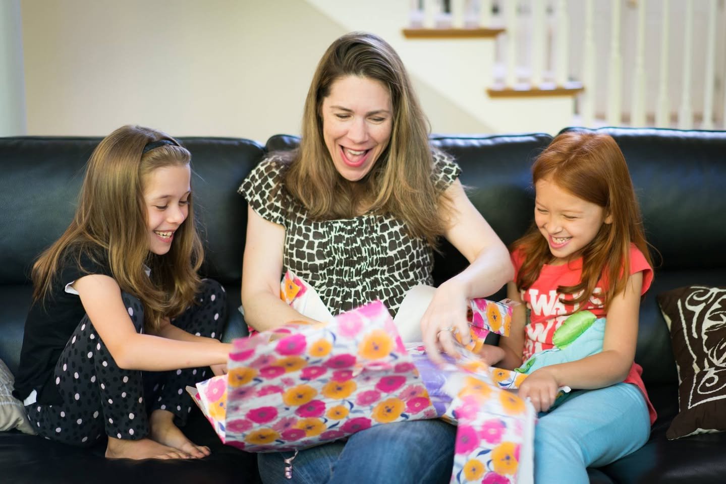 Happy Birthday, Kristie Three individuals enjoy wrapping colorful gifts together on a black leather couch, sharing smiles and laughter in a cozy home setting. The vibrant floral wrapping paper adds a festive touch to the heartwarming scene.