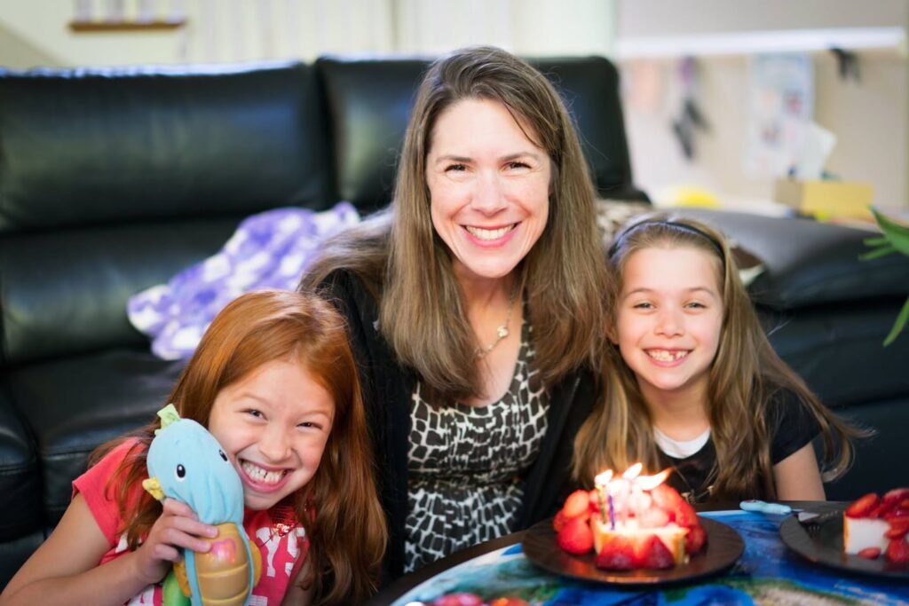 A woman and two girls smiling during a family celebration with a strawberry shortcake dessert.