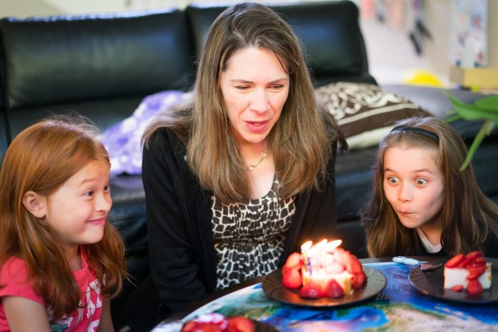A woman and two young girls gather around a table with strawberry-topped dessert plates. One girl is seen blowing out candles on a treat, while another smiles.