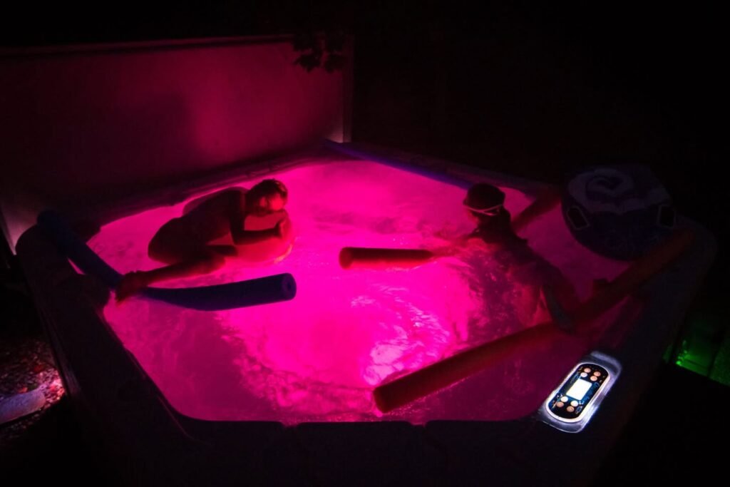 A dark scene showing a hot tub with magenta lighting and controls visible at the edge.