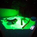 An outdoor swimming pool is illuminated with vibrant green night lighting. The image shows a person relaxing by the poolside.