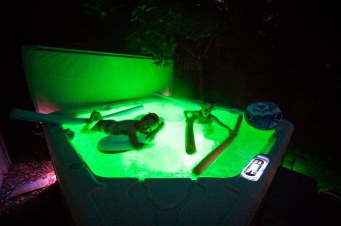 An outdoor swimming pool is illuminated with vibrant green night lighting. The image shows a person relaxing by the poolside.