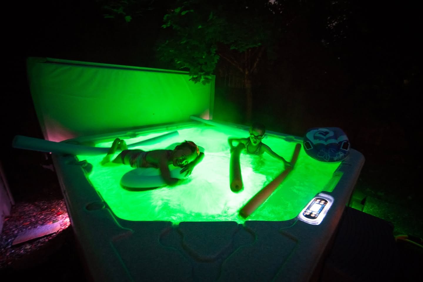 An outdoor swimming pool is illuminated with vibrant green night lighting. The image shows a person relaxing by the poolside.
