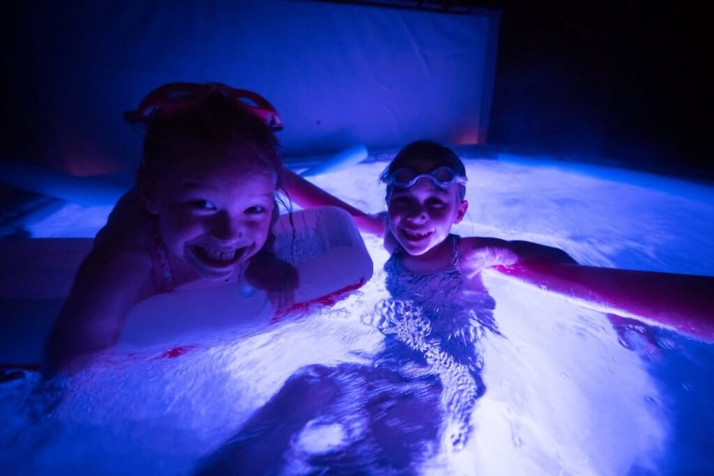 Two smiling people wearing goggles enjoy the glowing blue water of a night swim, capturing a moment of lighthearted activity.