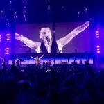 A concert scene featuring a performer with arms raised on stage under purple lights and a large screen with energetic audience silhouettes.