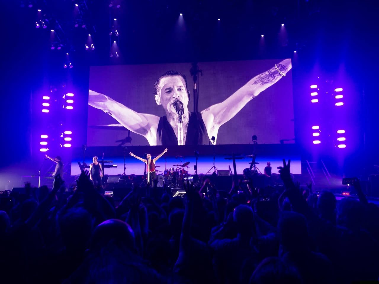 Depeche Mode: Spirit Tour A concert scene featuring a performer with arms raised on stage under purple lights and a large screen with energetic audience silhouettes.