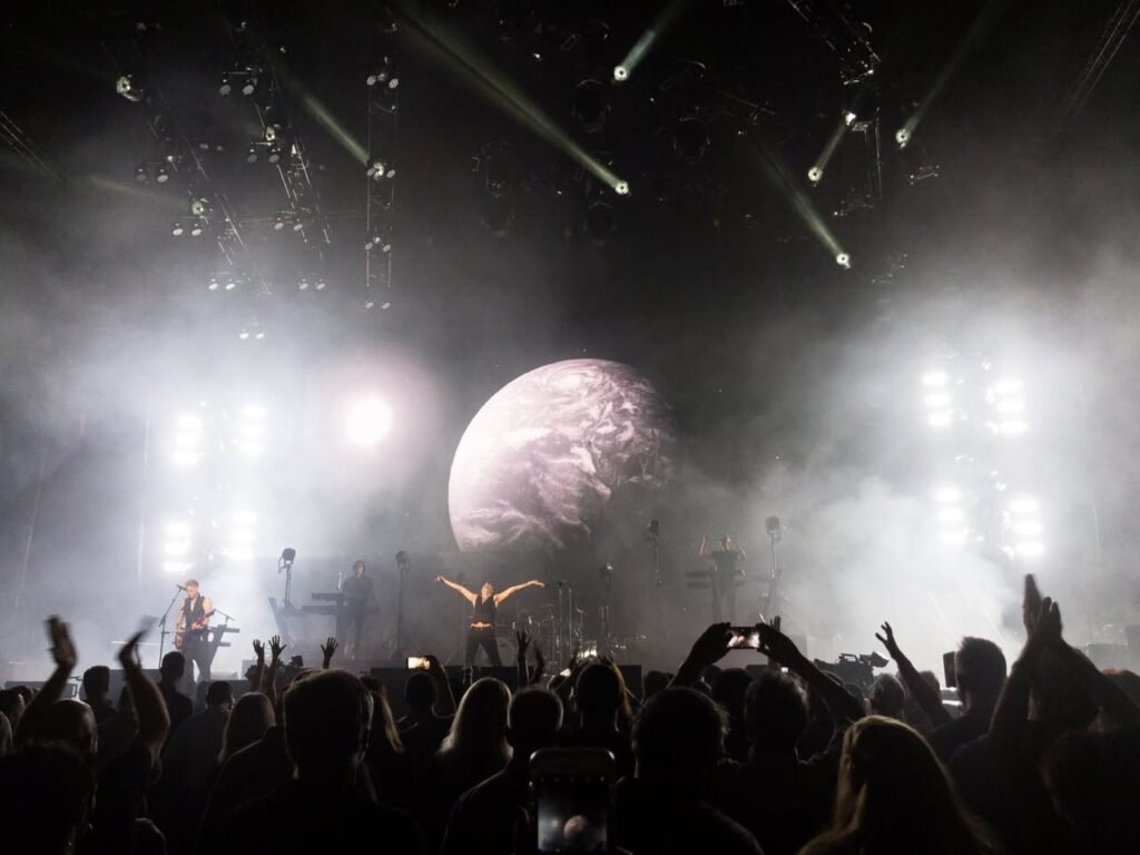 A dramatic concert scene featuring a large moon-like screen, stage lights, and an engaged audience.